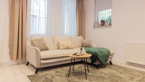 Ein Wohnzimmer mit einer Couch, einem kleinen runden Tisch mit einer Fernbedienung, einer grünen Tagesdecke und einem Fenster mit durchsichtigen Vorhängen.
