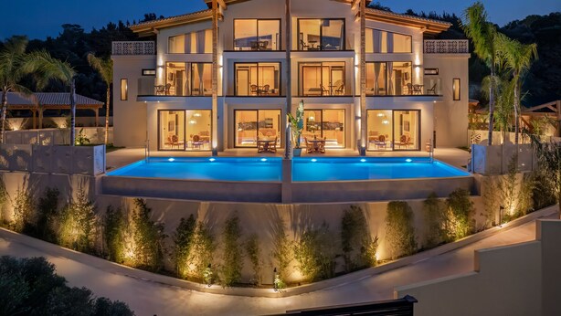 Una casa de varios pisos con piscina central, rodeada de jardines ajardinados y palmeras.