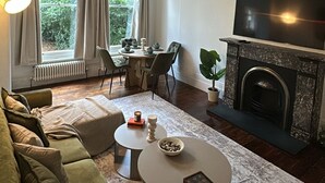 A living room with a sofa, coffee table, fireplace, TV, and large windows with curtains. - Cosy West Kensington Apartment - 3 Min to Tube (London)