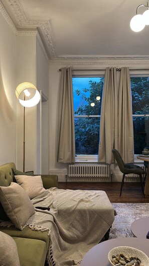 A cozy room with a green sofa, a patterned throw blanket, a side table with a bowl, and a chair. - Cosy West Kensington Apartment - 3 Min to Tube (London)
