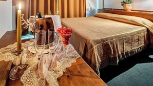 A neatly made bed with a checkered bedspread, a bedside table with a candle, glasses, and a bowl of fruit. - Suite in Hotel Cristallino (Cortina d'Ampezzo)
