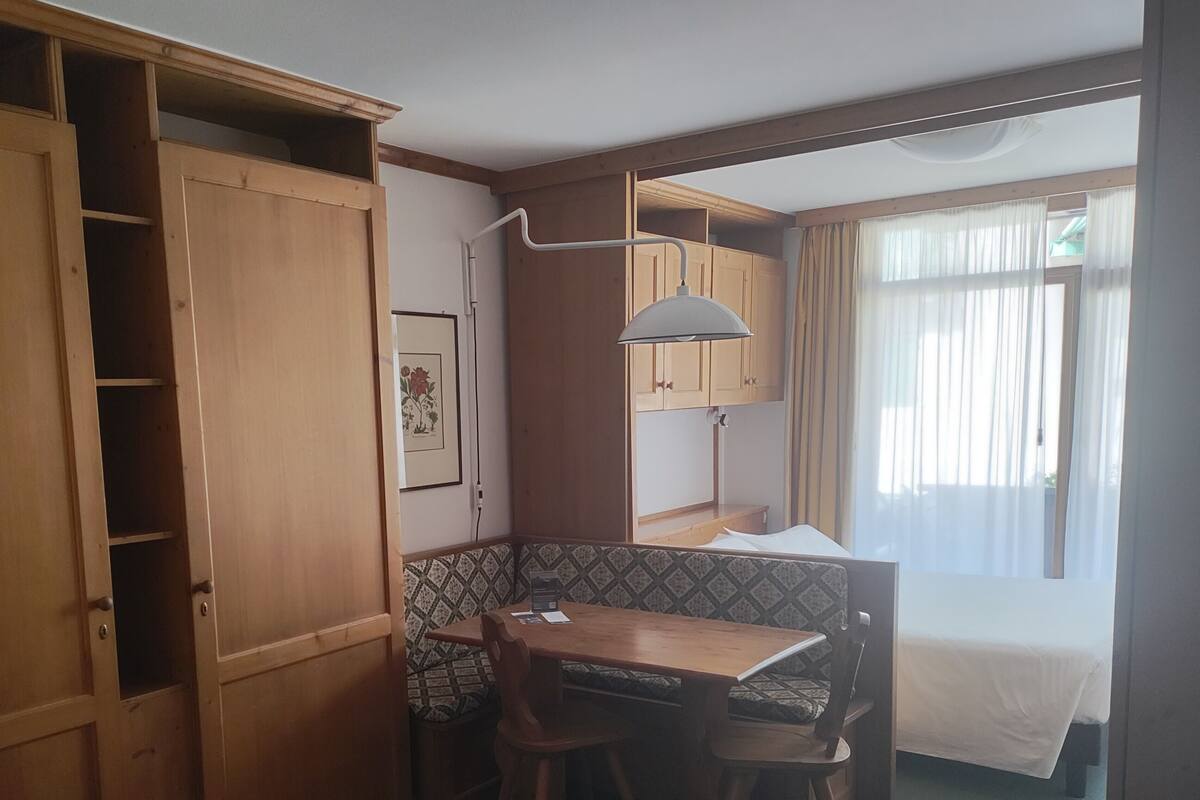 Ein gemütliches Zimmer mit Bett, Schreibtisch und Stuhl. Ein Fenster mit Vorhängen, eine Wandleuchte und eine Tür mit Ablagefläche.