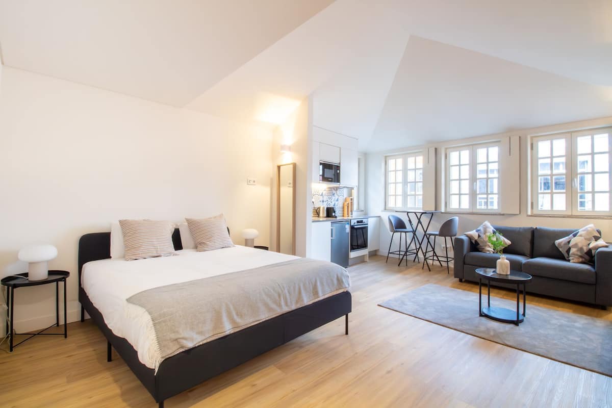 Un dormitorio moderno con una cama grande, una kitchenette pequeña y una zona de estar con sofá y sillones.