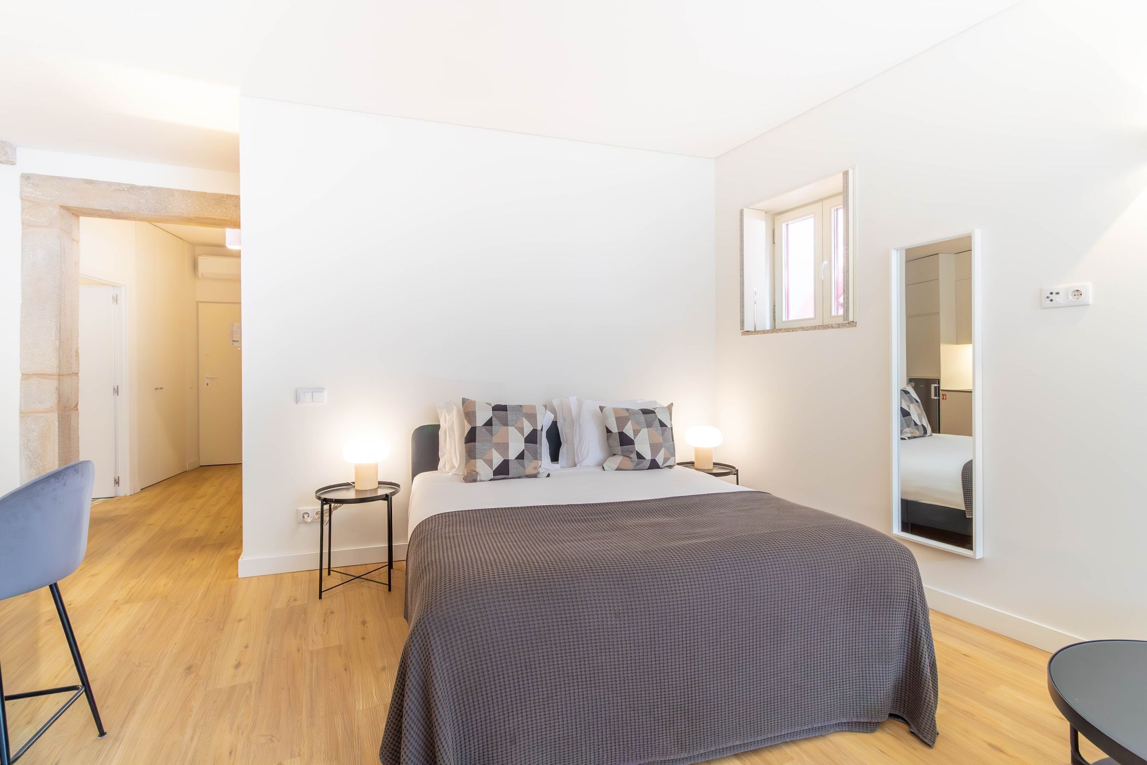 Un dormitorio con cama, mesitas de noche, una silla, un espejo y una puerta que da a otra habitación.