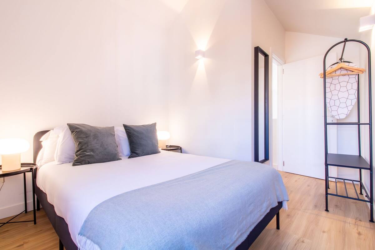Una cama bien hecha con edredón blanco y gris, una mesita de noche con lámpara y un estante de pared con un objeto de diseño geométrico.