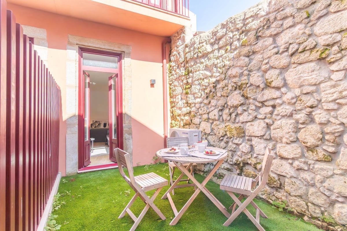 Un pequeño patio con una mesa, sillas, un muro de piedra y una puerta que da a un espacio interior.