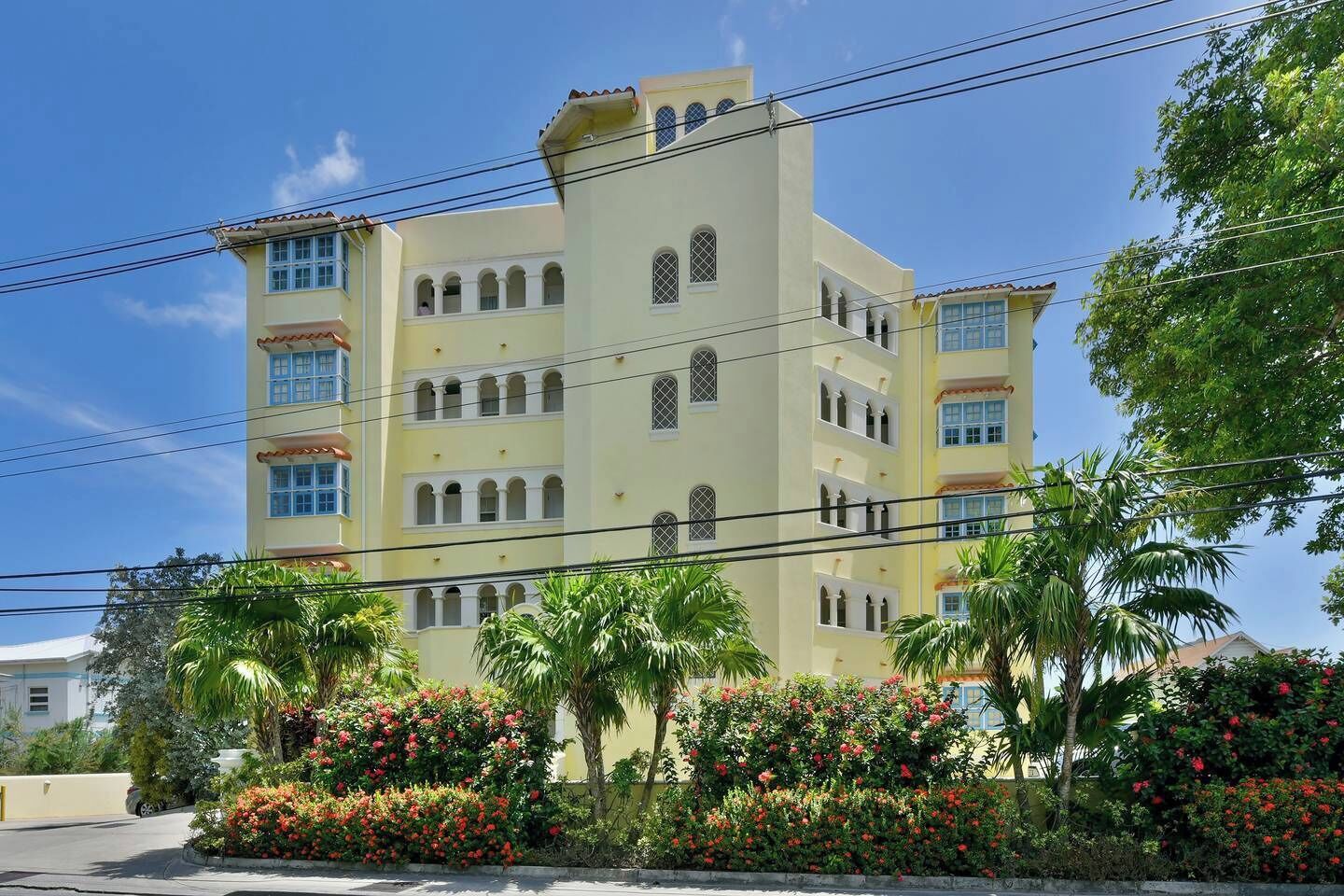 A multi-story hotel with a light yellow facade, multiple balconies, and a garden with red flowers.