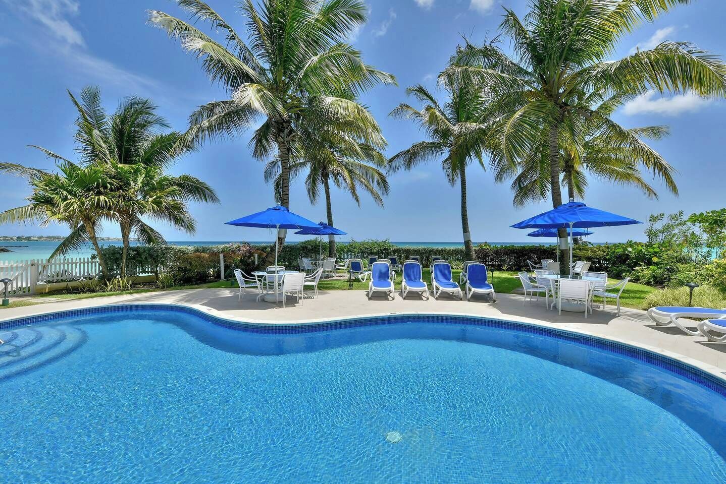 A pool area with lounge chairs and umbrellas, surrounded by palm trees and a view of the ocean.