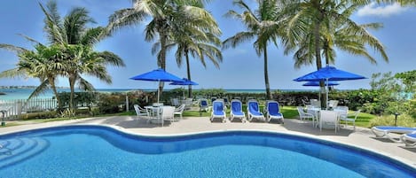A pool area with lounge chairs and umbrellas, surrounded by palm trees and a view of the ocean.