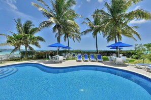 Zona de piscina con sillones de playa y sombrillas, rodeada de palmeras y vista al océano.