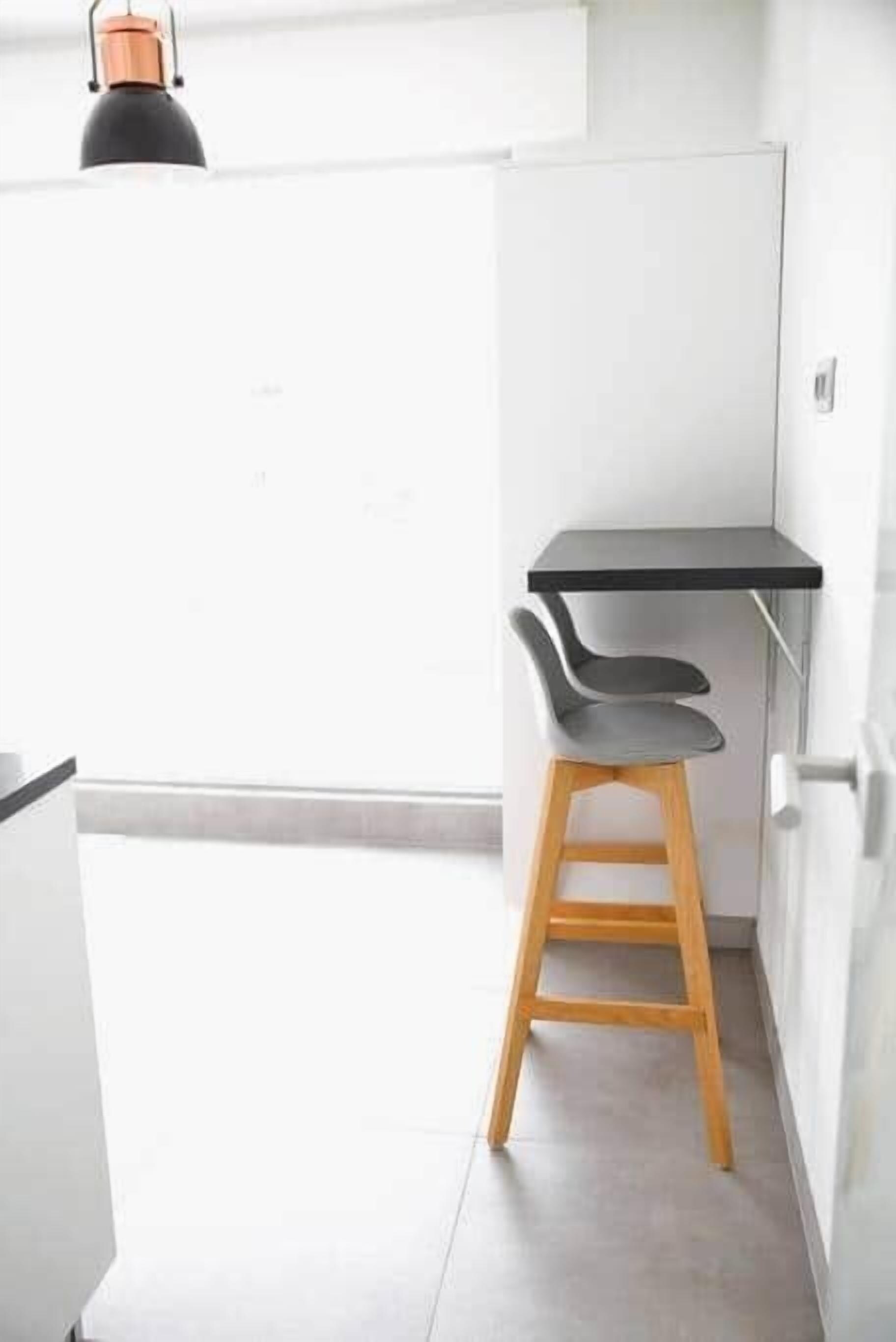 Une cuisine moderne avec un plan de travail blanc, un tabouret en bois et une suspension noire.
