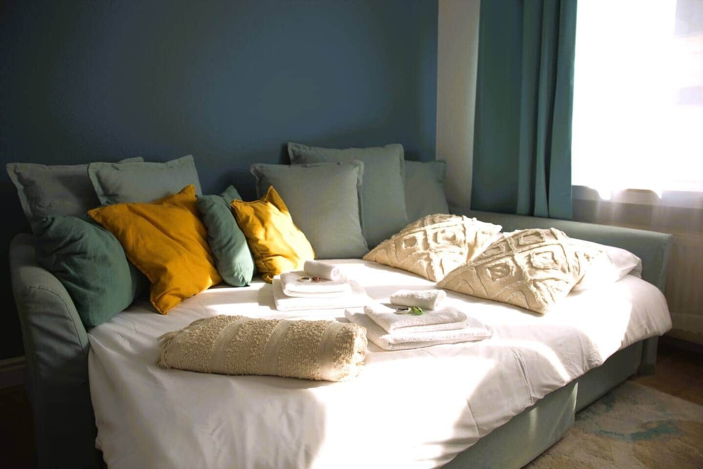 A bed with white linens, multiple pillows in various colors, and a folded towel on the bed.