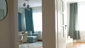 A spacious living area with a light-colored wooden floor, a white sofa, a white chair, and a small white table. There is a window with blue curtains and a hanging light fixture. - Beautiful Cosy 2BR Apartment (Luxembourg City)