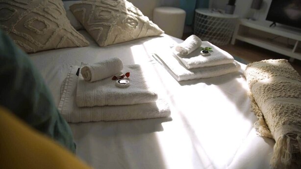 A neatly made bed with folded towels and a rolled-up towel, a small round tray with a bottle, and a white blanket on the right side. - Beautiful Cosy 2BR Apartment (Luxembourg City)