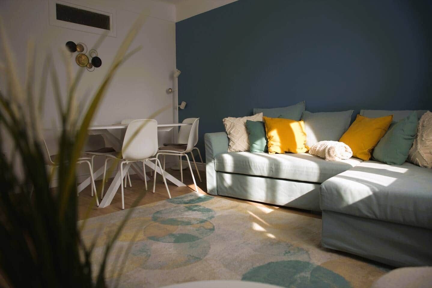 A living room with a sofa, a dining table with chairs, and a plant in the foreground.