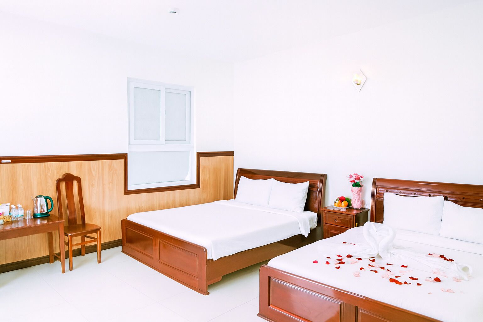 A hotel room with two beds, a wooden desk, a chair, and a window.