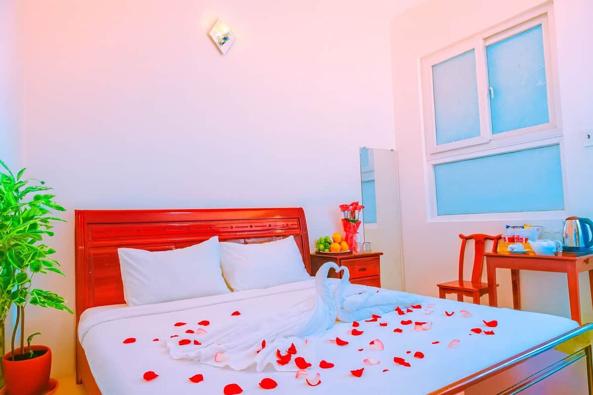 A neatly made bed with white linens and red rose petals scattered on it. There is a red headboard, a bedside table with a vase of flowers, and a small table with a kettle and cups.