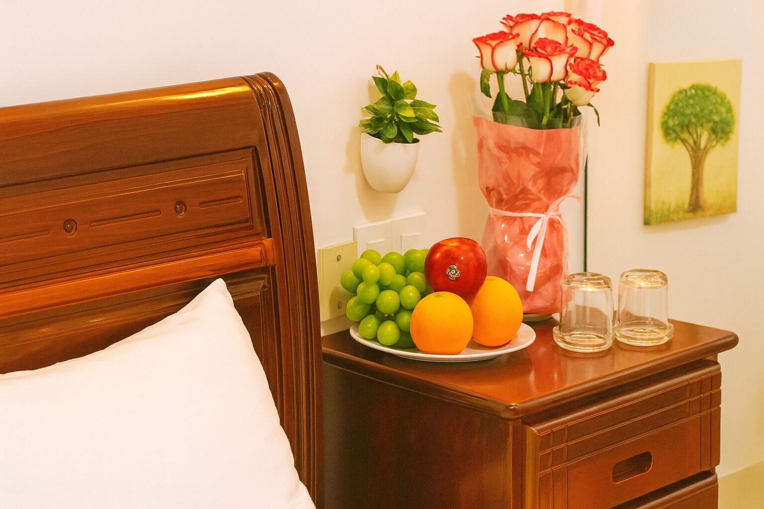 A neatly made bed with a white pillow, a wooden nightstand with a plate of fruit, a vase with red roses, a small potted plant, and a framed picture on the wall.