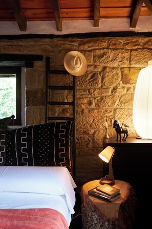 A cozy room with a stone wall, a wooden ceiling, a bed with white and red bedding, a nightstand with a lamp, a hat, and a small statue. - Molino Tejada: Apartamento Neruda (Polientes)
