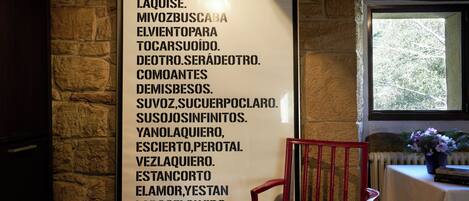 Un poème encadré est accroché à un mur de pierre, avec une fenêtre et une chaise au premier plan.