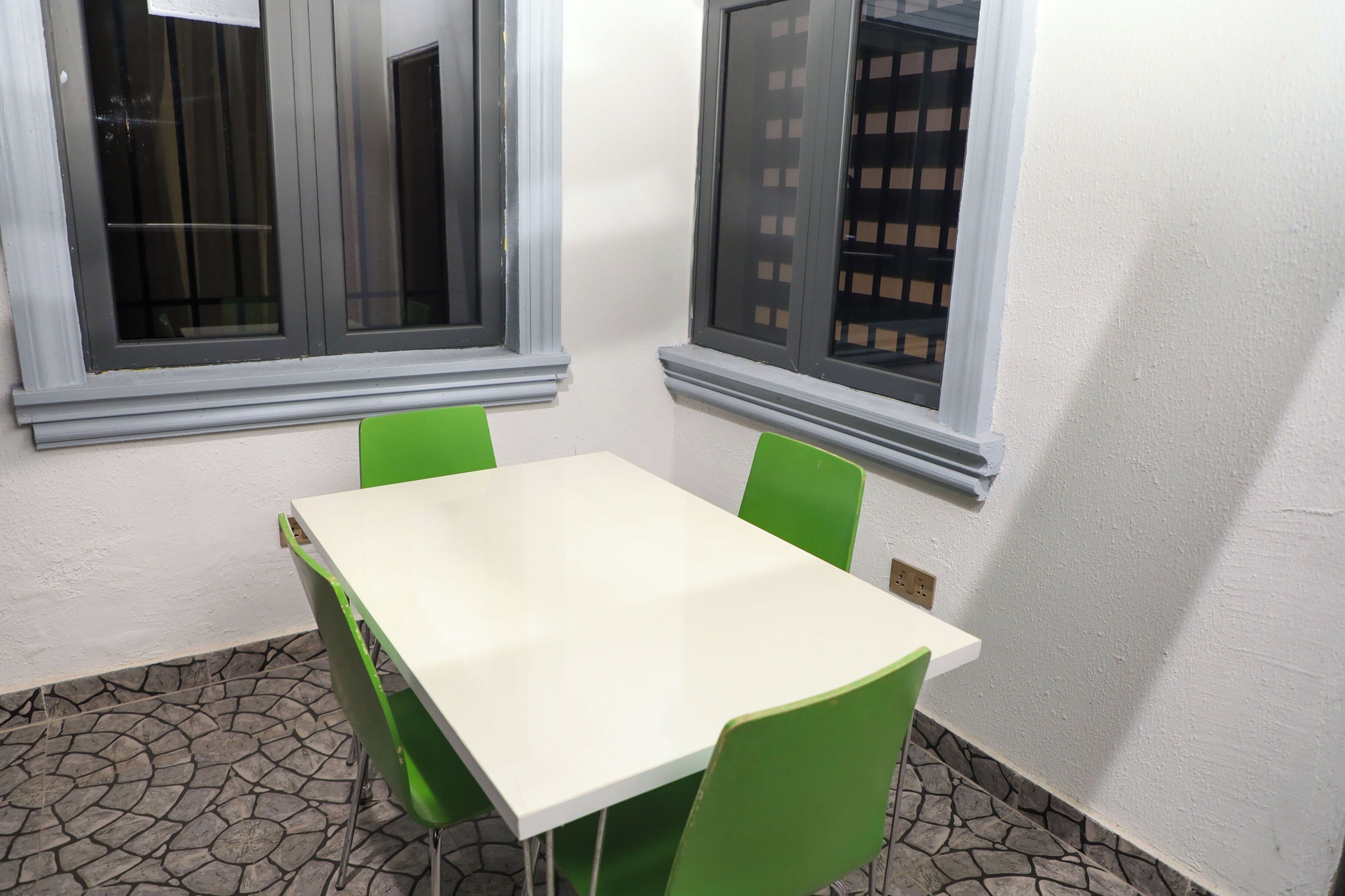 Ein kleiner Raum mit einem weißen Tisch, grünen Stühlen, einem Fenster mit Metallgitter und einem bodenähnlichen Steinmuster.