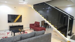 A modern living room with a grey sofa, red armchair, and a black coffee table. A staircase with glass railings leads to an upper level. - ED - Havilah Suites (Abuja)