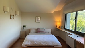 Een slaapkamer met een houten bed, een nachtkastje, een potplant en een raam met uitzicht op bomen.