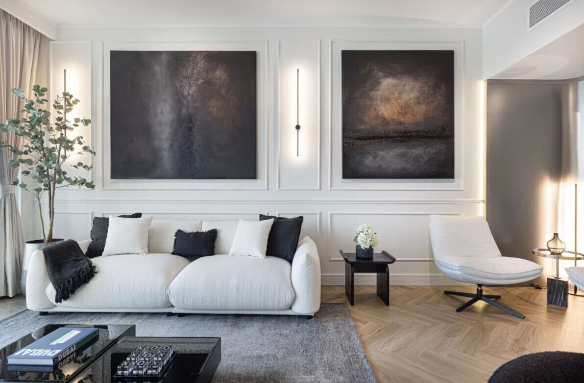 Uma sala de estar moderna com um sofá branco, uma mesa lateral com um vaso, e uma poltrona. Há duas grandes pinturas abstratas na parede.