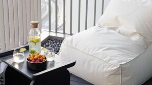 En balkon med en hvid polstret stol, en bakke med vandflaske, glas og en skål med frugt.