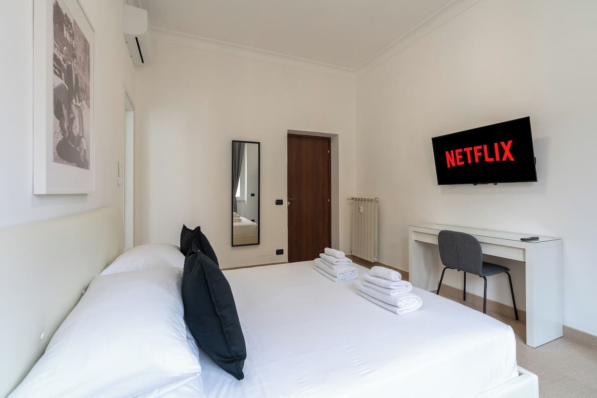 A hotel room with a bed, a desk, a chair, a TV displaying "NETFLIX", a door, and a radiator.