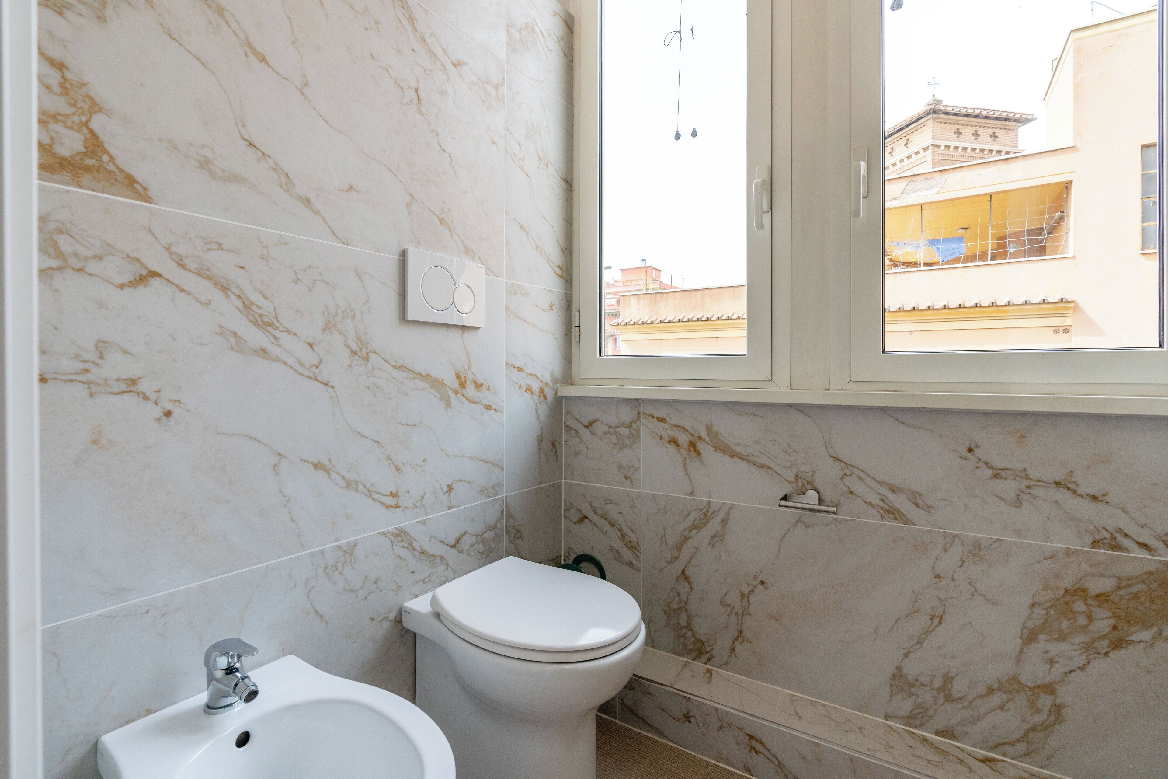 Een badkamer met toilet, bidet en raam met uitzicht op gebouwen.
