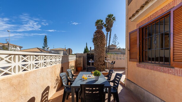 Un patio al aire libre con una mesa y sillas, rodeado por una cerca blanca, palmeras y un edificio con una ventana.