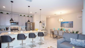 A modern kitchen with white cabinets, a central island with bar stools, stainless steel appliances, pendant lighting, and a dining area with a gray sofa and a painting on the wall. - Marvelous 4 Bd w Pool Waterpark Near Disney 218 (Kissimmee)