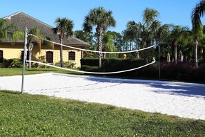 Ein Volleyballfeld mit Netz, umgeben von Sand und Grünflächen, mit einem Gebäude im Hintergrund.