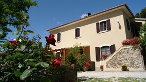 Egy kétszintes épület, melyhez kőből készült lépcső vezet a bejárathoz, és amelyet buja zöld növényzet és vörös virágok öveznek.