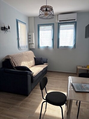 A small, modern living room with a sofa, a dining table, and two windows with blue frames. - Cottage in St Palais with 2 beds + a baby, garden, terrace, quiet (Saint-Palais-sur-Mer)