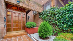 A brick building with a large wooden door, a small garden area with shrubs, and a tiled pathway leading to the entrance. - P Casa Cafe Madrid Calle Carlos Dubois 3 (Madrid)