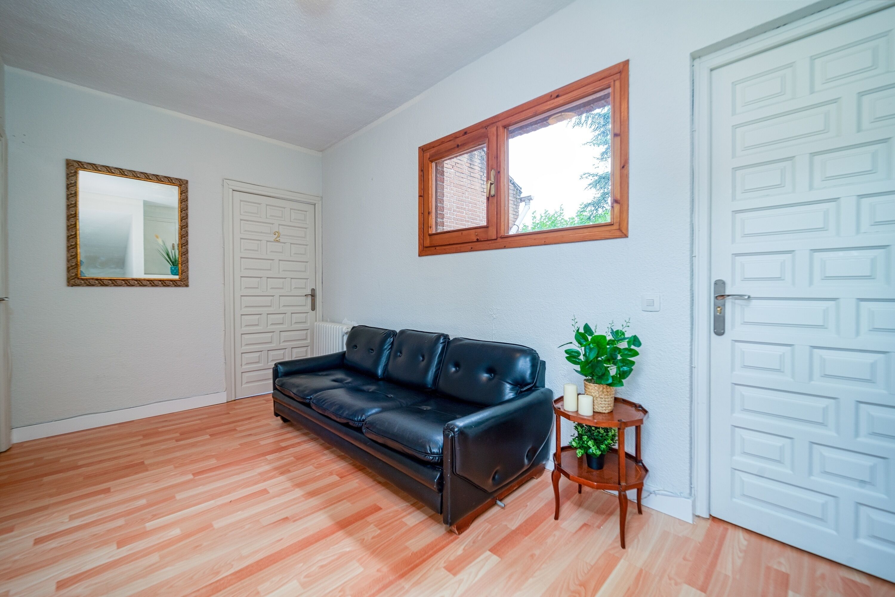 Una habitación con un sofá de cuero negro, una mesita de madera con una planta, un espejo y una ventana con marco de madera.