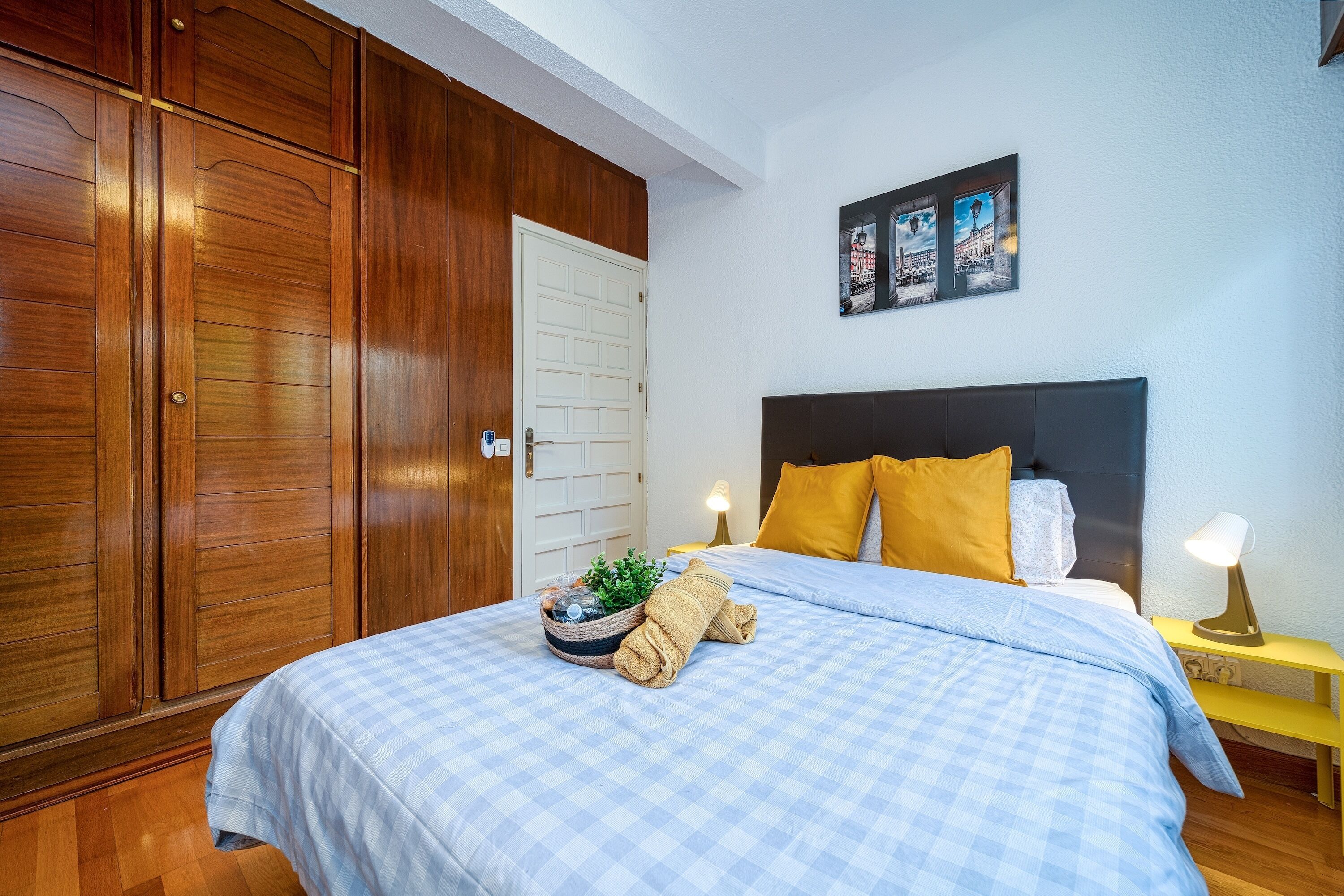 Une chambre avec une grande armoire, un lit recouvert d’une courtepointe à carreaux, une table de chevet avec une lampe et un mur orné de tableaux encadrés.