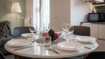 Uma mesa de jantar posta com pratos, copos e uma fruteira, em um quarto com um sofá e micro-ondas ao fundo.