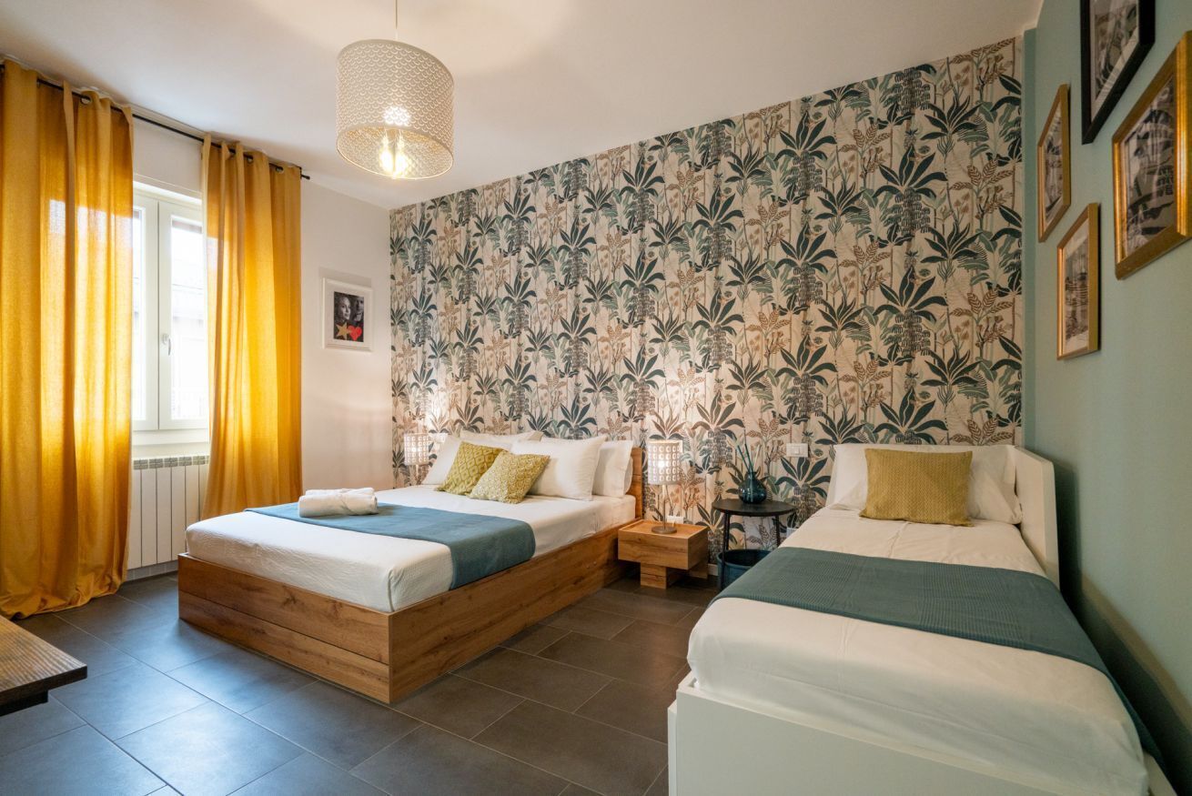 Una camera da letto con due letti, una finestra con tende gialle, carta da parati decorata e un lampadario.