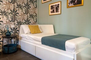 A single-bed room with a white bed, a blue blanket, a yellow pillow, a small round table with a vase, and a blue pet bed.