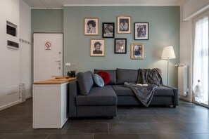 Un salon avec un canapé gris, un petit meuble blanc et une machine à café. Des tableaux encadrés ornent le mur, et une lampe se trouve sur la droite.