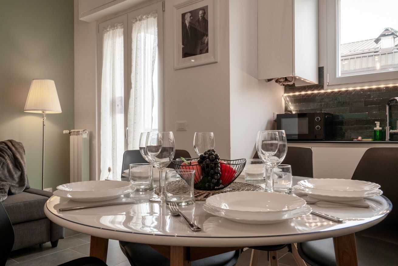 Un'area pranzo con un tavolo rotondo apparecchiato per quattro persone, una ciotola di frutta e una cucina sullo sfondo.