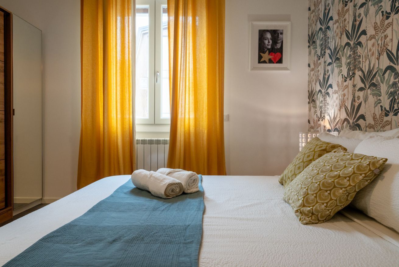 Ein Hotelzimmer mit einem Bett, einer blauen Decke, zwei Kissen, einem gerahmten Bild an der Wand und gelben Vorhängen.