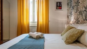 Ein Hotelzimmer mit einem Bett, einer blauen Decke, zwei Kissen, einem gerahmten Bild an der Wand und gelben Vorhängen.