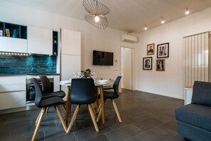 Eine moderne Küche mit blauer Rückwand, ein Essbereich mit rundem Tisch und Stühlen, ein an der Wand befestigter Flachbildfernseher und ein graues Sofa im Wohnbereich.