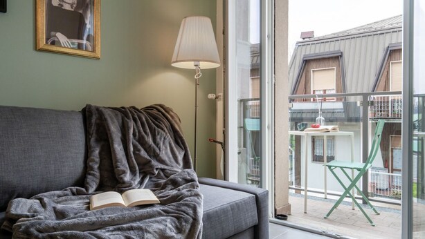 Et værelse med en grå sofa, en åben bog, en gulvlampe og udsigt til en balkon med stol og bord.