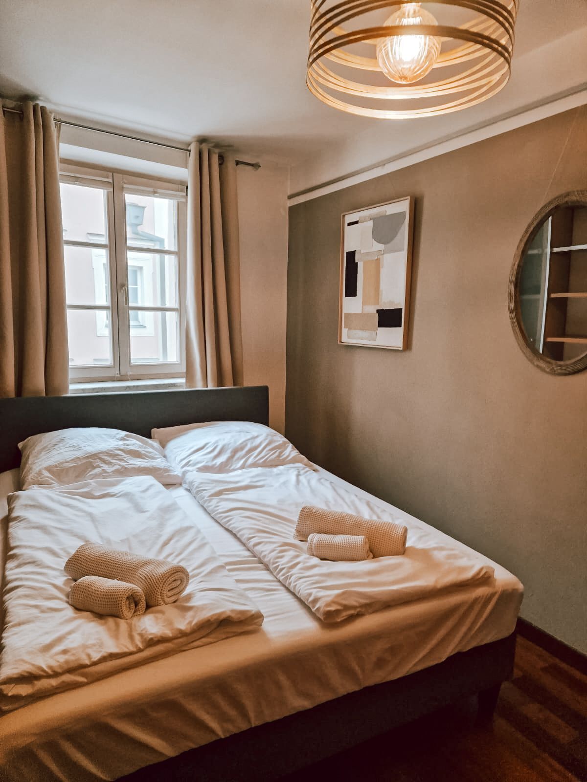 A neatly made bed with two rolled towels, a window with curtains, a wall-mounted abstract art piece, and a round mirror.