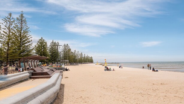 Ein Strand mit einer Holzpromenade, Sitzbereichen und hohen Bäumen.